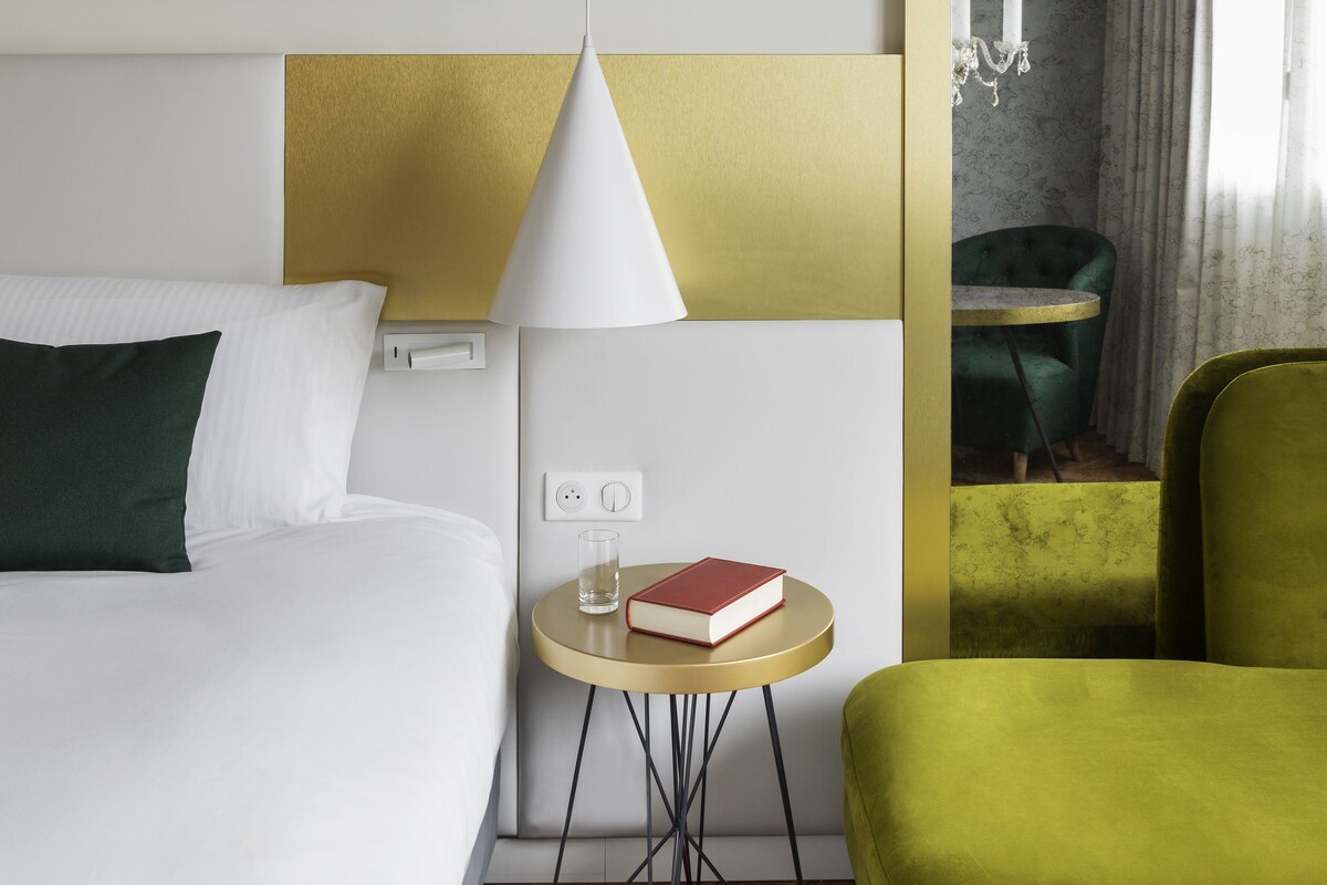 Une chambre d’hôtel avec un lit, une petite table ronde sur laquelle se trouvent un livre et un verre, un fauteuil vert et un miroir qui reflète un fauteuil vert et des rideaux.