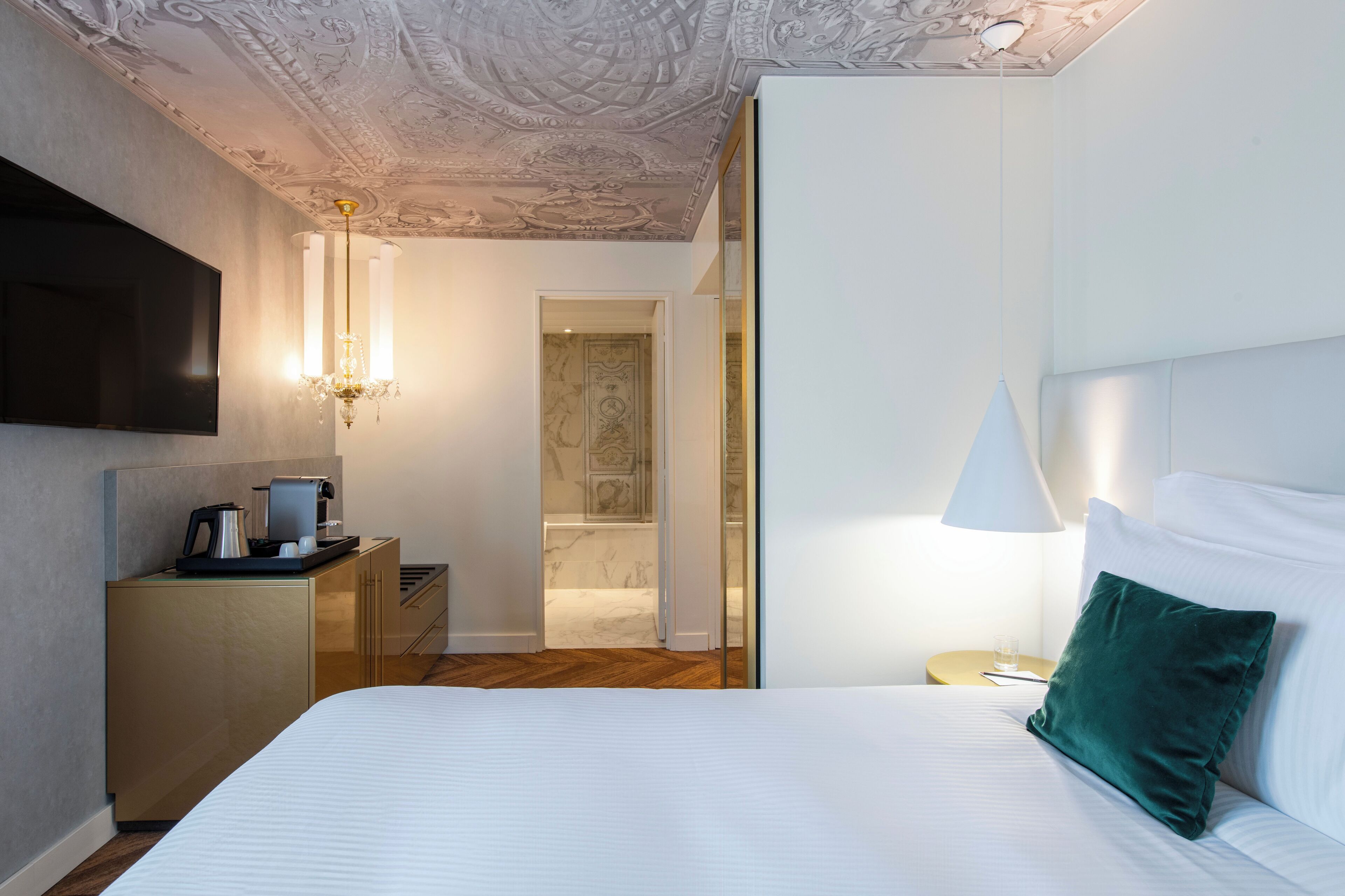 Una habitación de hotel con cama, televisión, cafetera, lámpara y puerta que da al baño.
