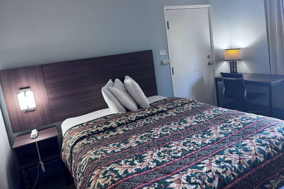 A hotel room with a bed, a nightstand with a phone, a desk with a lamp, and a door.