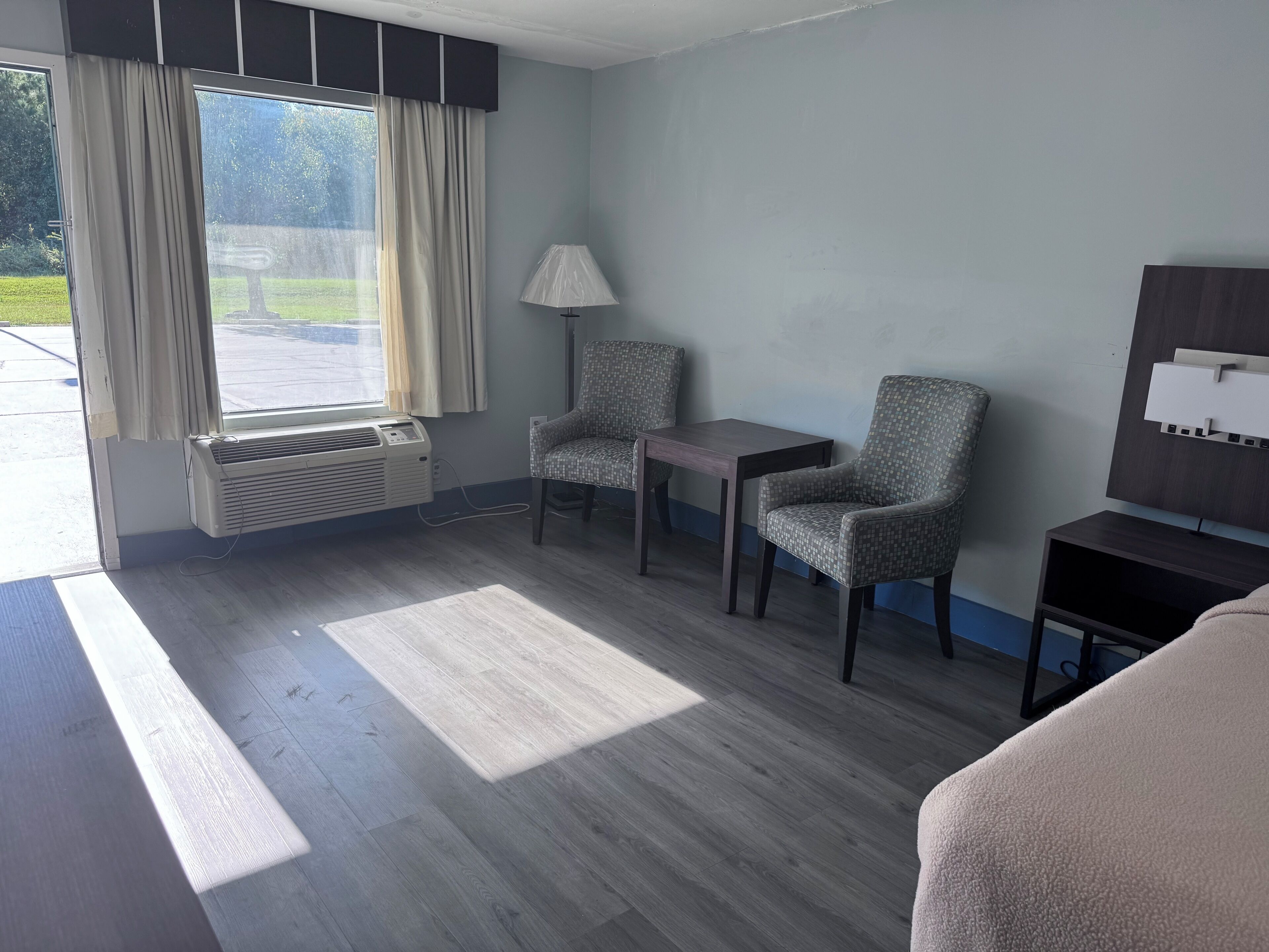 Habitación de hotel con un ventanal grande, un calefactor portátil, dos sillones, una mesita, una lámpara y un televisor sobre un mueble de madera.