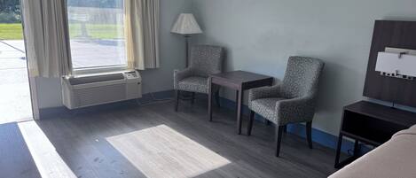 Habitación de hotel con un ventanal grande, un calefactor portátil, dos sillones, una mesita, una lámpara y un televisor sobre un mueble de madera.