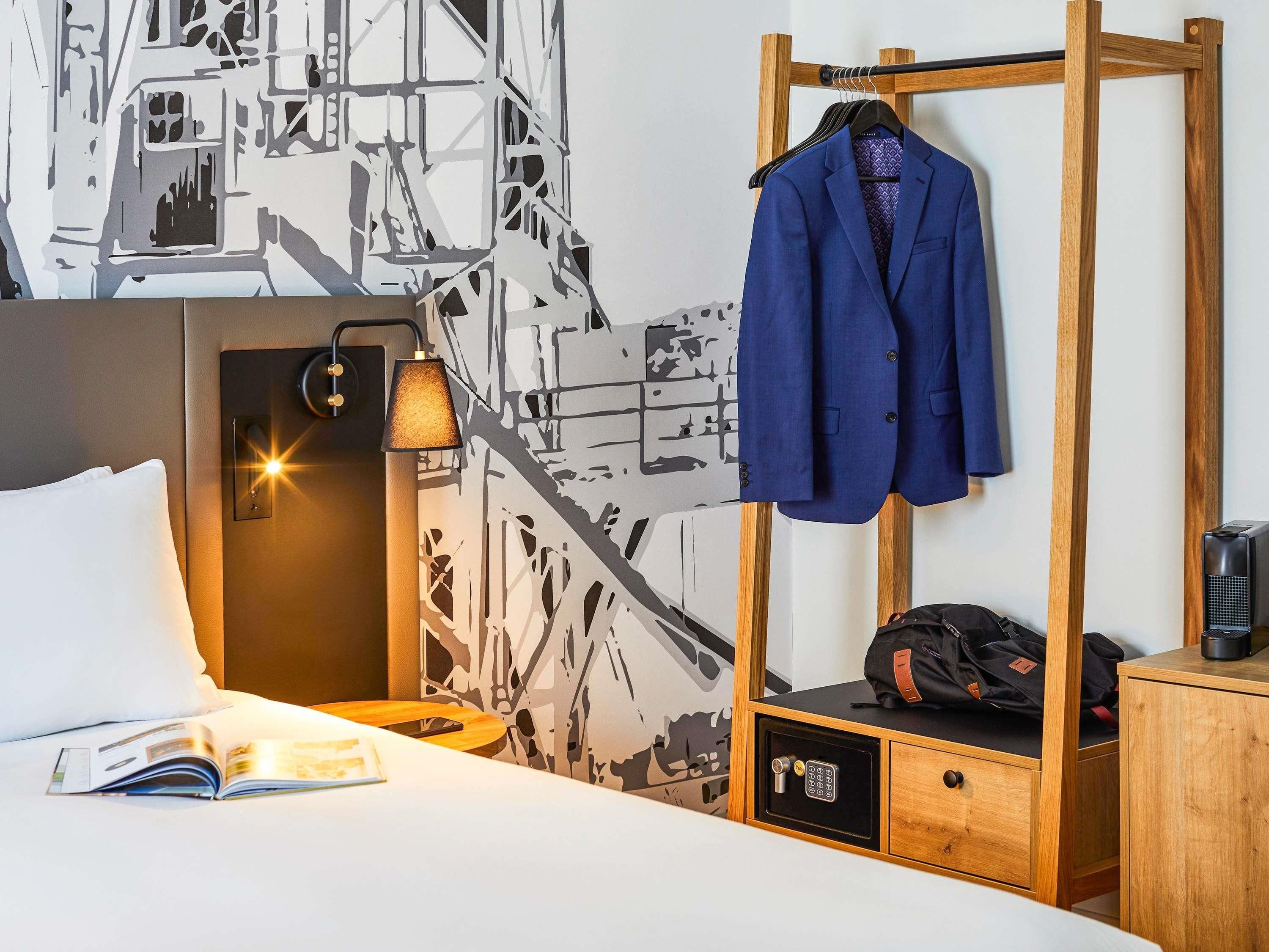 Ein Hotelzimmer mit Bett, Kleiderhaken, Nachttisch mit Lampe und einer Wand mit Stadtbild-Wandmalerei.