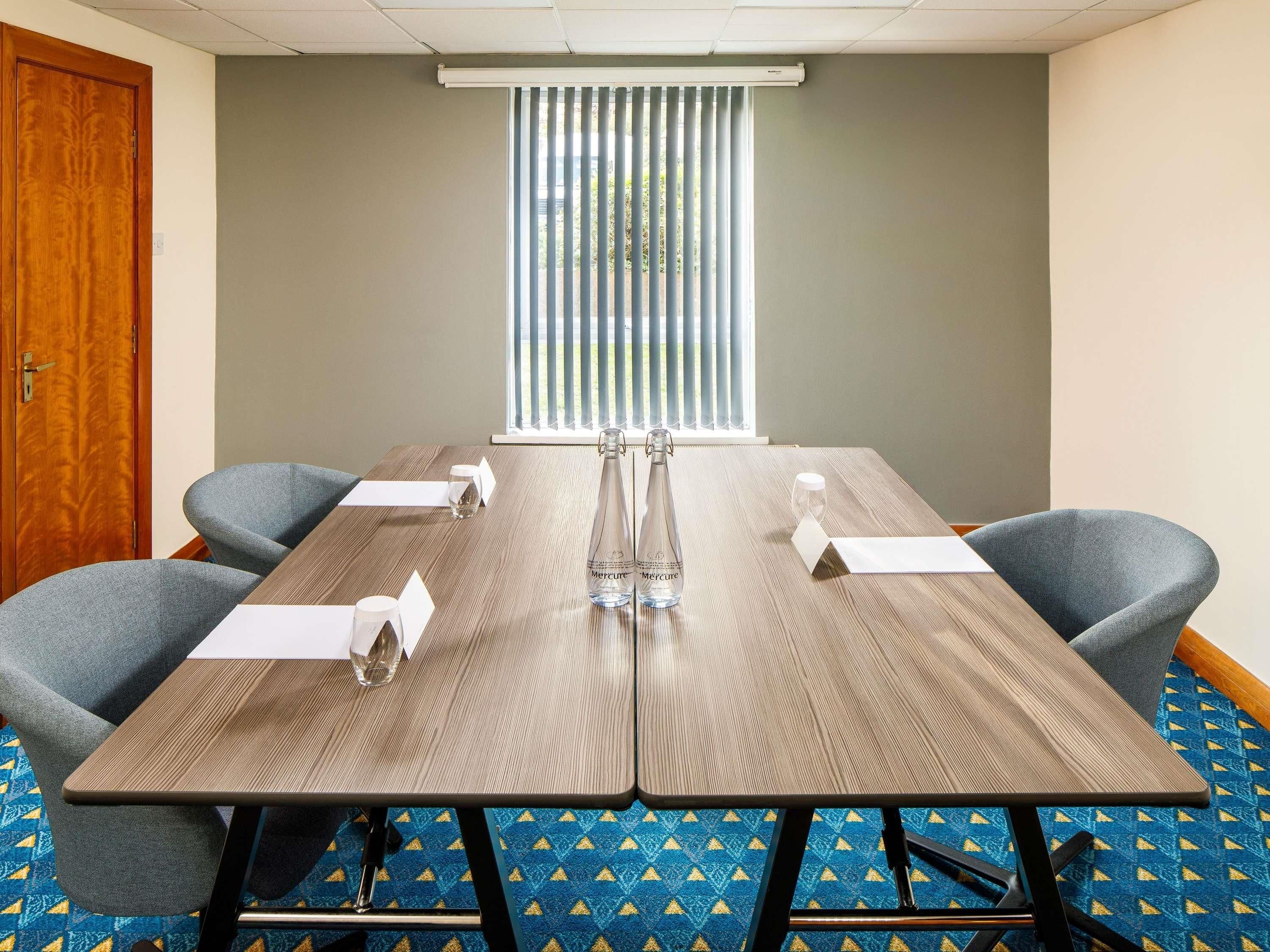 A conference room with a wooden table, four grey chairs, a bottle of water, and a window with blinds.