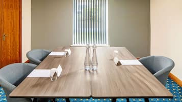 Un salón de conferencias con una mesa de madera, cuatro sillas grises, una botella de agua y una ventana con persianas.