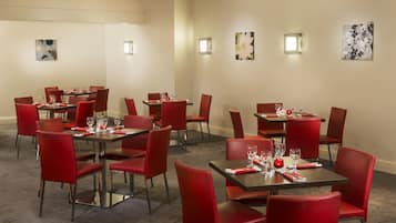 A hotel dining area with multiple tables and red chairs, equipped with glassware and napkins.