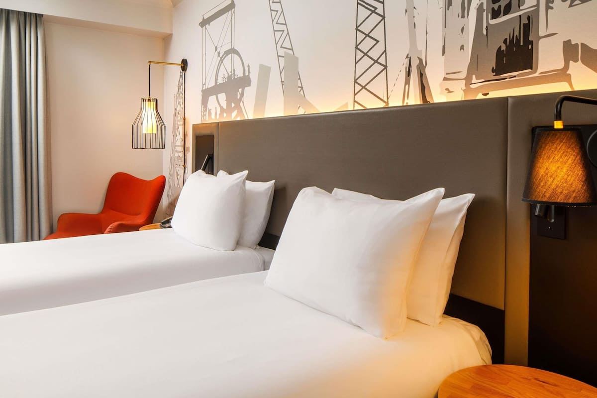 A hotel room with two beds, a wooden side table, a red chair, and a wall with industrial-themed artwork.