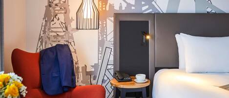 A hotel room with a bed, a red chair, a small table with a phone and cup, and a wall with a cityscape mural.