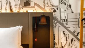 Une chambre d’hôtel avec un lit, une petite table ronde en bois, une lampe fixée au mur et un papier peint à motifs.