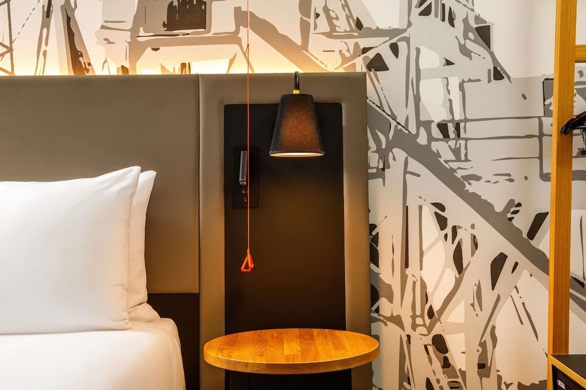 A hotel room with a bed, a small round wooden table, a wall-mounted lamp, and a patterned wall.