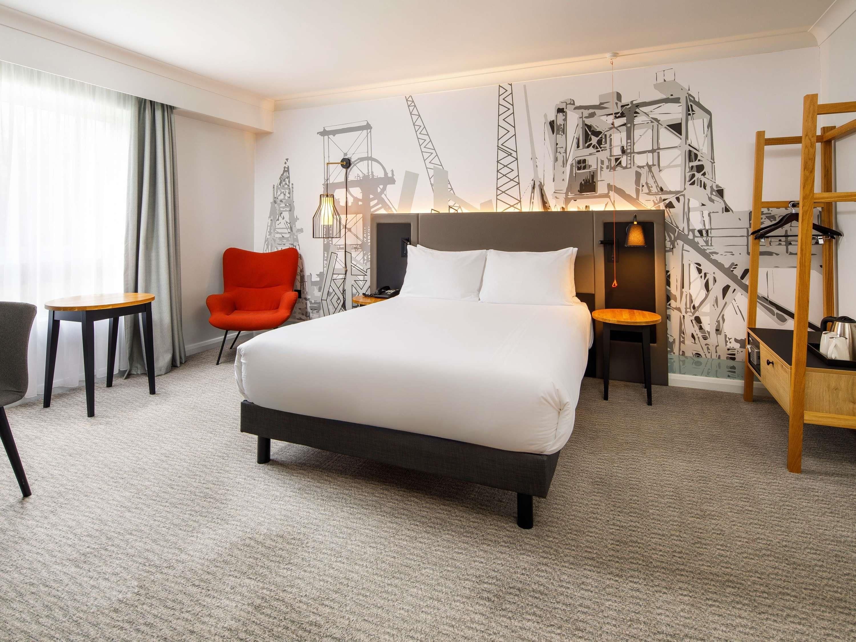 A hotel room with a large bed, a red chair, a small table, and a wall with construction-themed artwork.