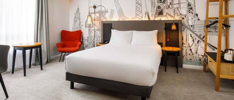 A hotel room with a large bed, a red chair, a small table, and a wall with construction-themed artwork.