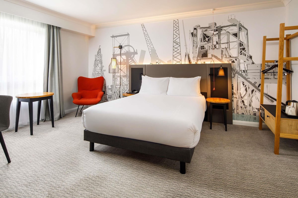 A hotel room with a large bed, a red chair, a small table, and a wall with construction-themed artwork.