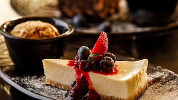Prato de sobremesa com uma fatia de cheesecake coberta com frutas vermelhas e morango, acompanhado de uma tigela que parece conter um doce ou pudim.
