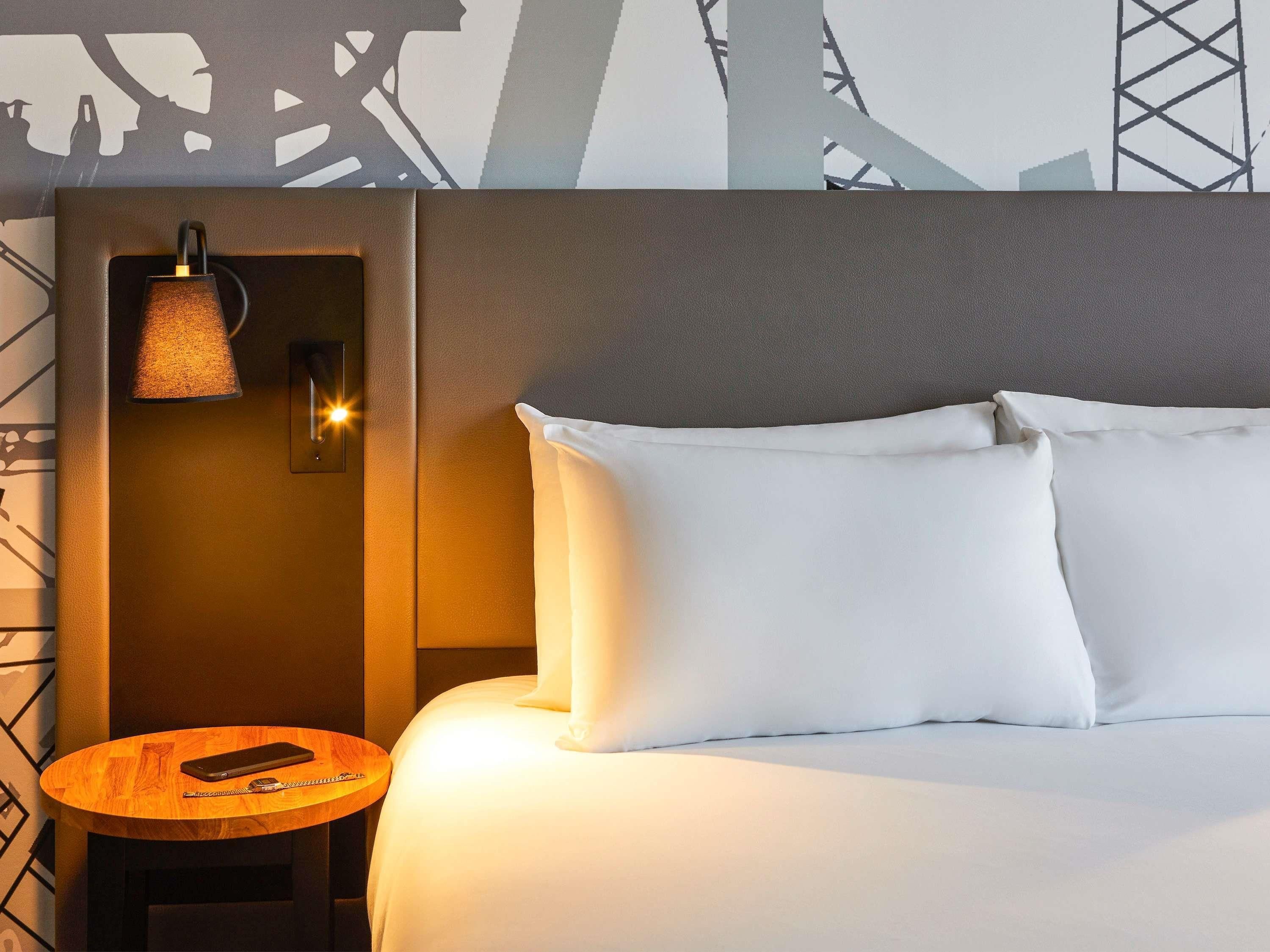 Habitación de hotel con cama, almohadas, mesita de noche con teléfono y una lámpara de pared.