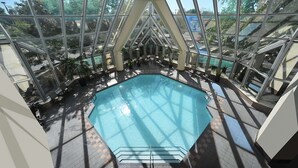 Une piscine intérieure dotée d’un toit en verre, entourée de chaises longues et de plantes en pot.