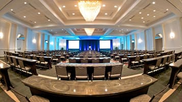A large conference room with rows of chairs and tables, a stage with a blue backdrop, and a chandelier.