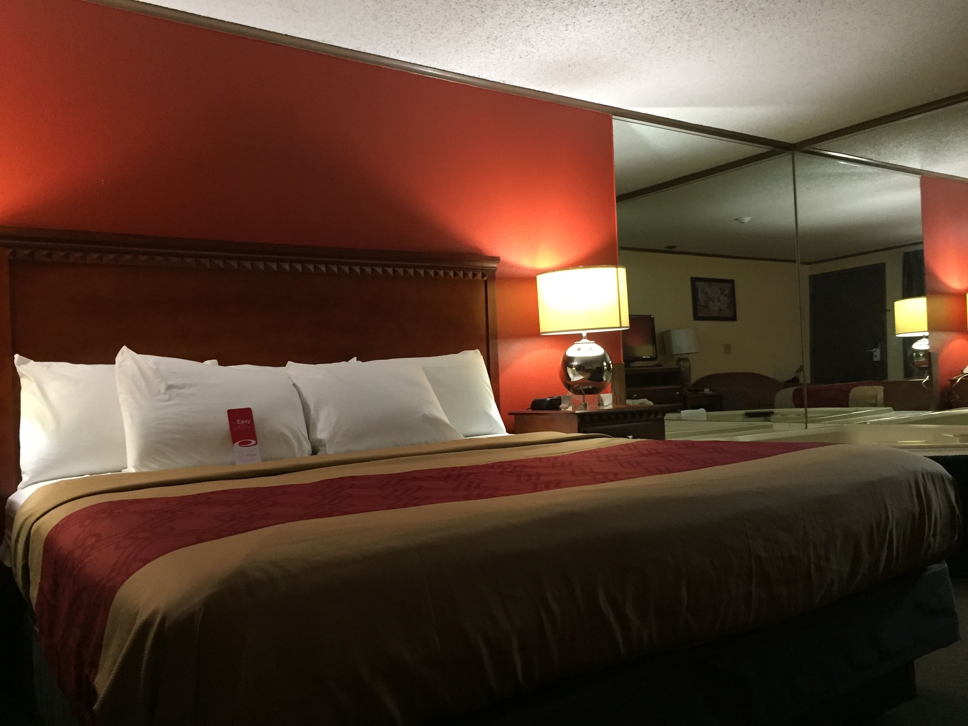 Une chambre d’hôtel avec un grand lit, une tête de lit en bois, un mur rouge et un miroir sur le mur opposé.