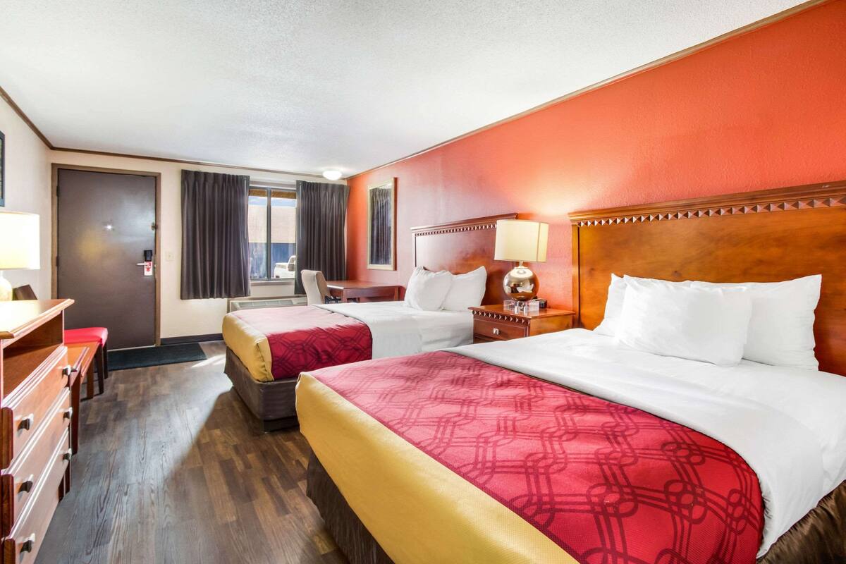A hotel room with two beds, a wooden headboard, a desk with a chair, and a window with curtains.