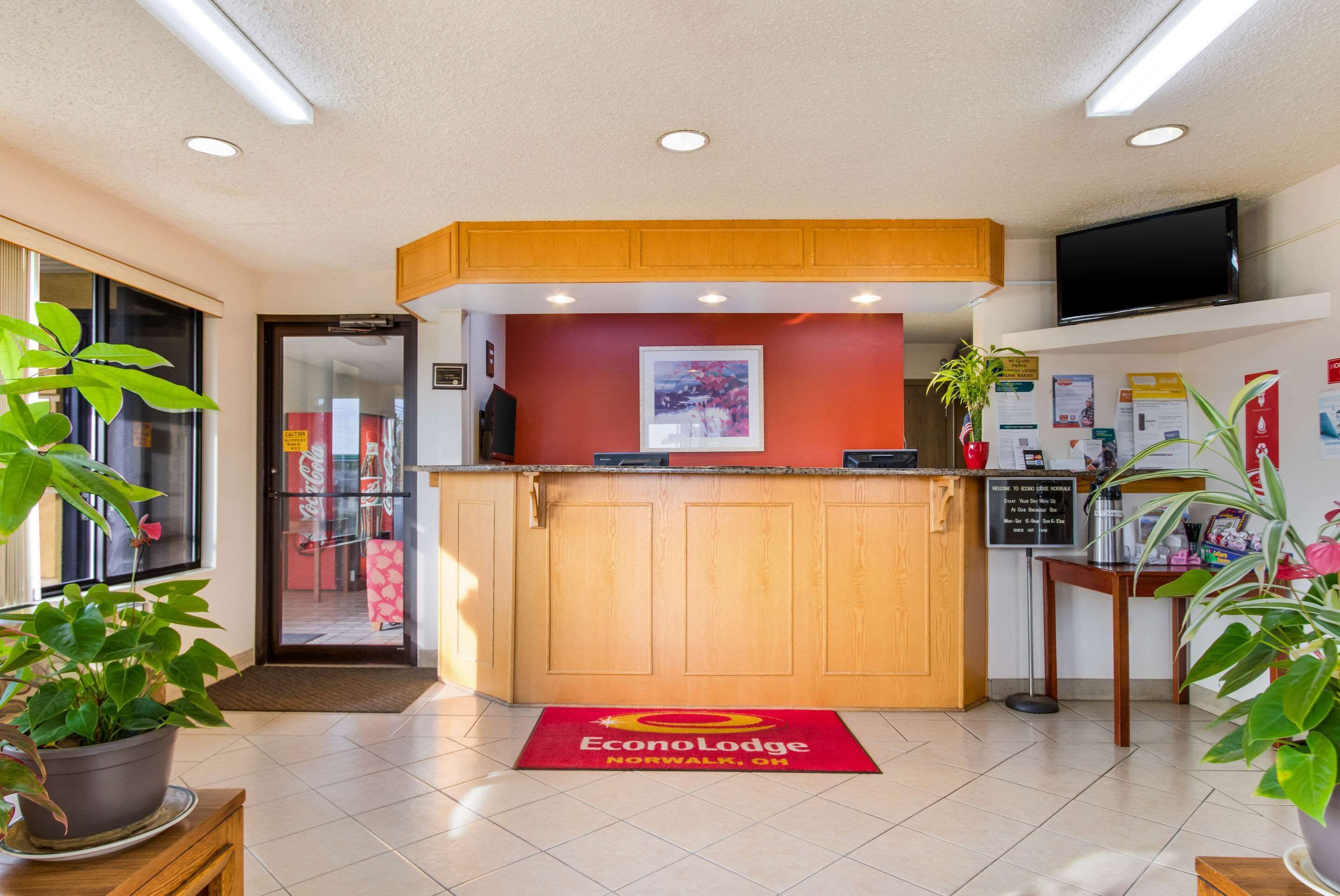 A reception area with a wooden counter, a red carpet with the hotel name, a television on the wall, and informational posters.