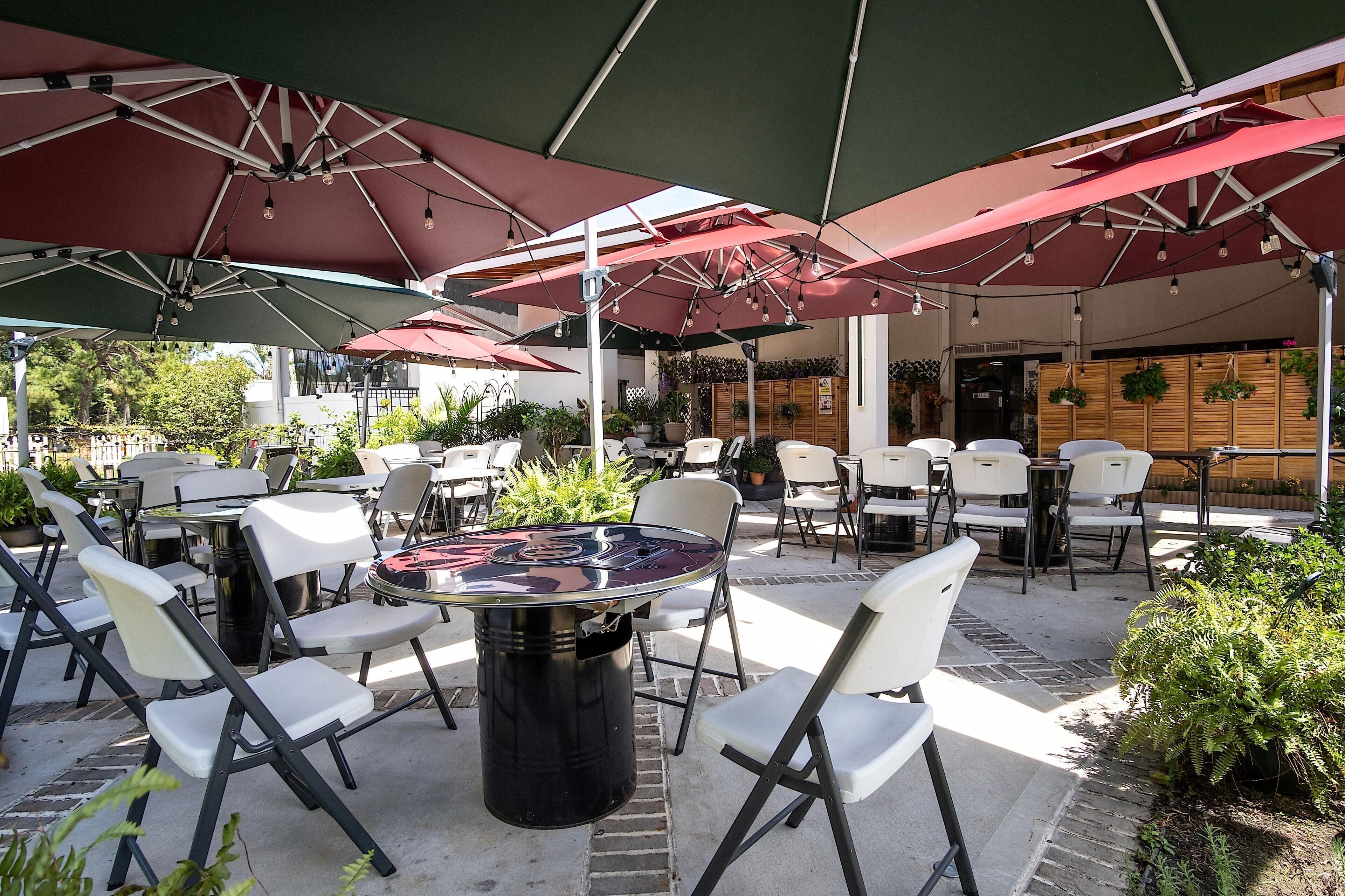 An outdoor patio with tables and chairs under large umbrellas, surrounded by potted plants and a wooden wall.