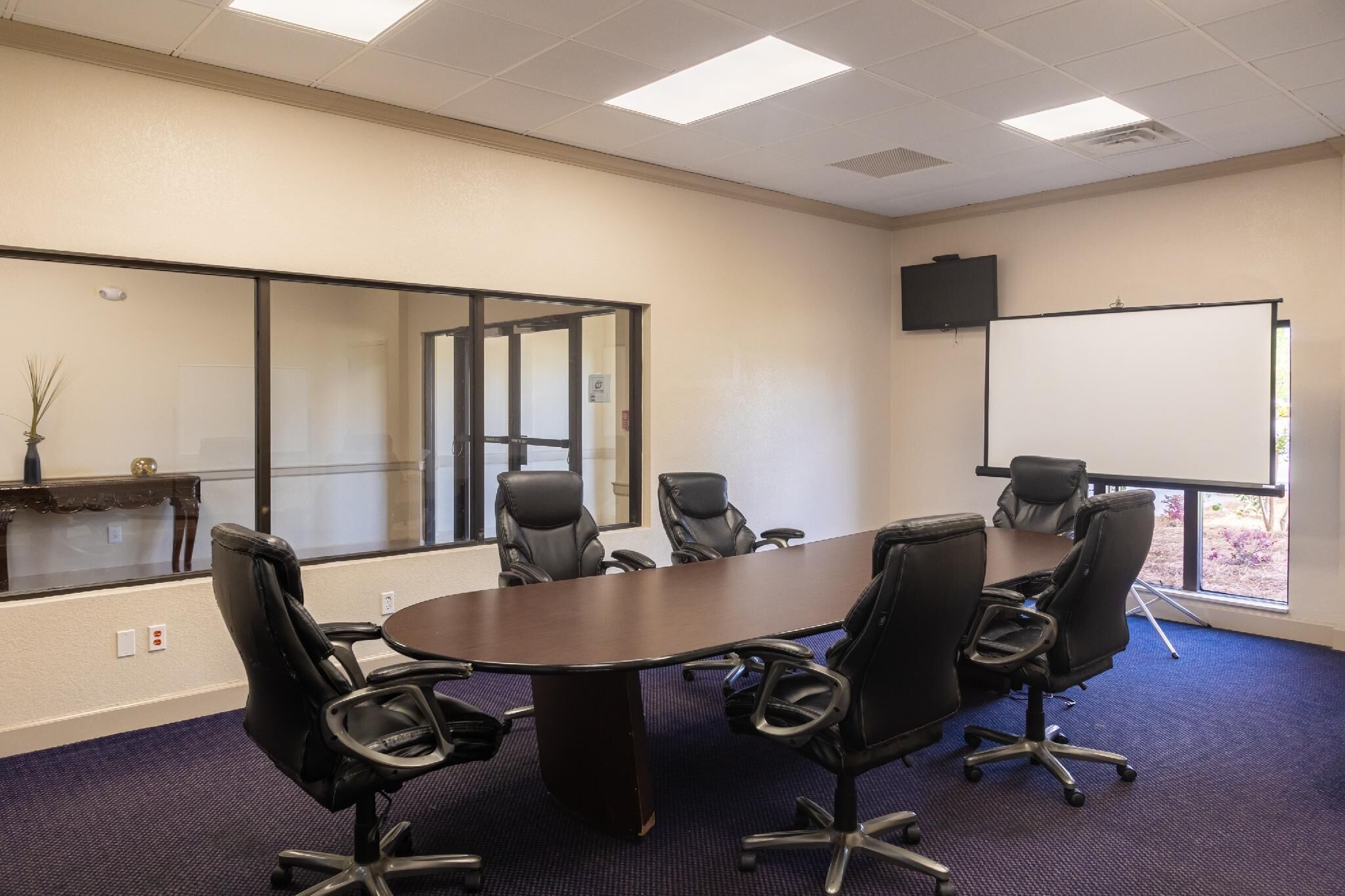 A conference room with a large table, six black chairs, a whiteboard, and a television on the wall.