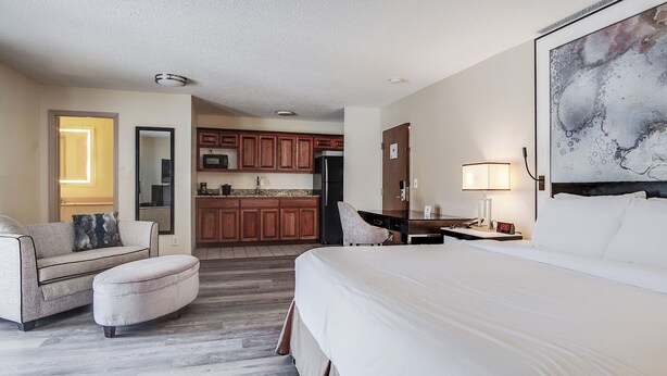 A hotel room with a large bed, a sofa, a small table, a chair, a desk, a lamp, and a kitchen area in the background. - Cottonwood Suites Savannah Hotel & Conference Center (Pooler)