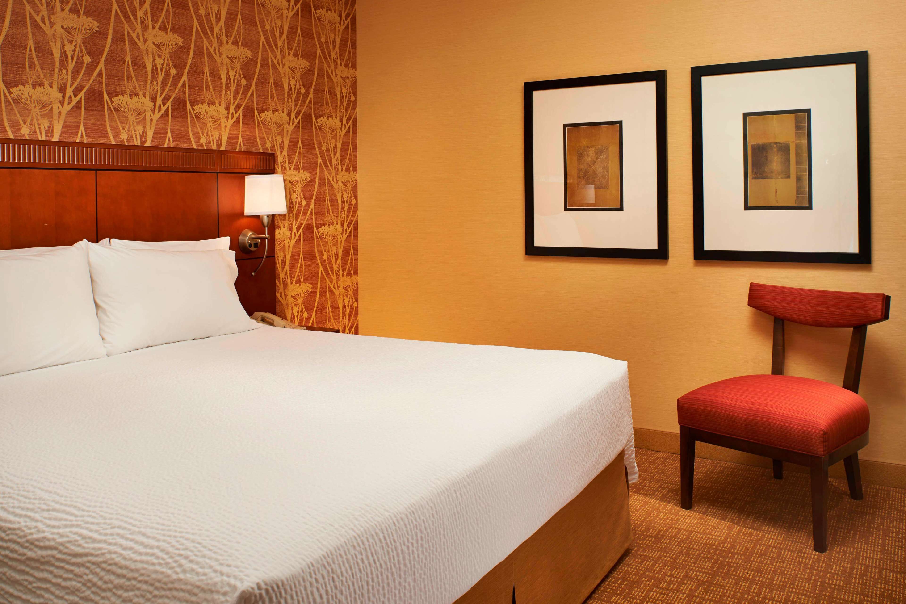 A hotel room with a large bed, a bedside table with a lamp, a chair, and two framed pictures on the wall.