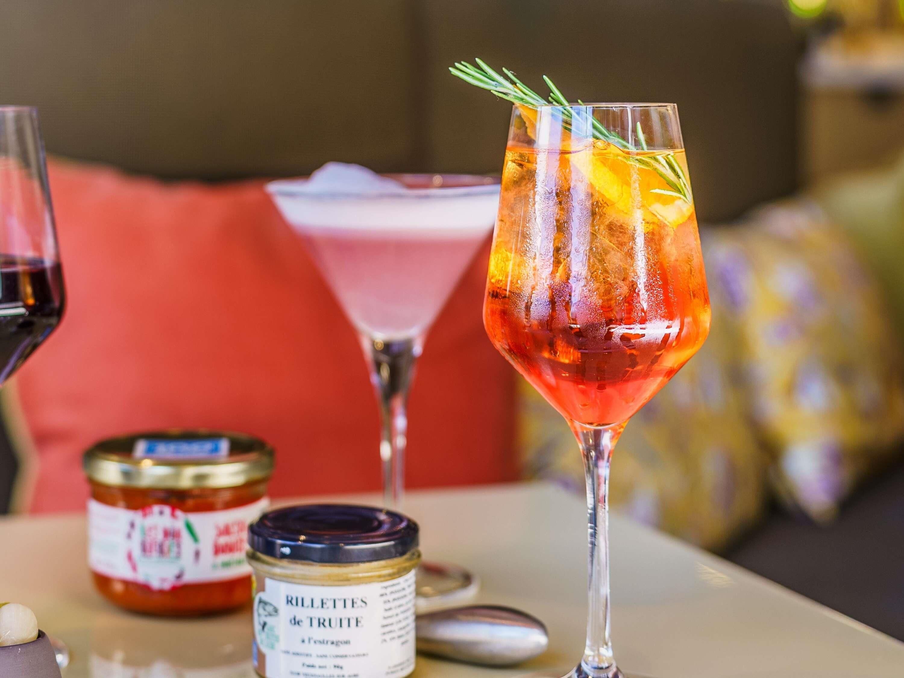 Una mesa con un vaso de bebida roja decorada con una ramita de romero, un frasco de mermelada y una botella de vino.