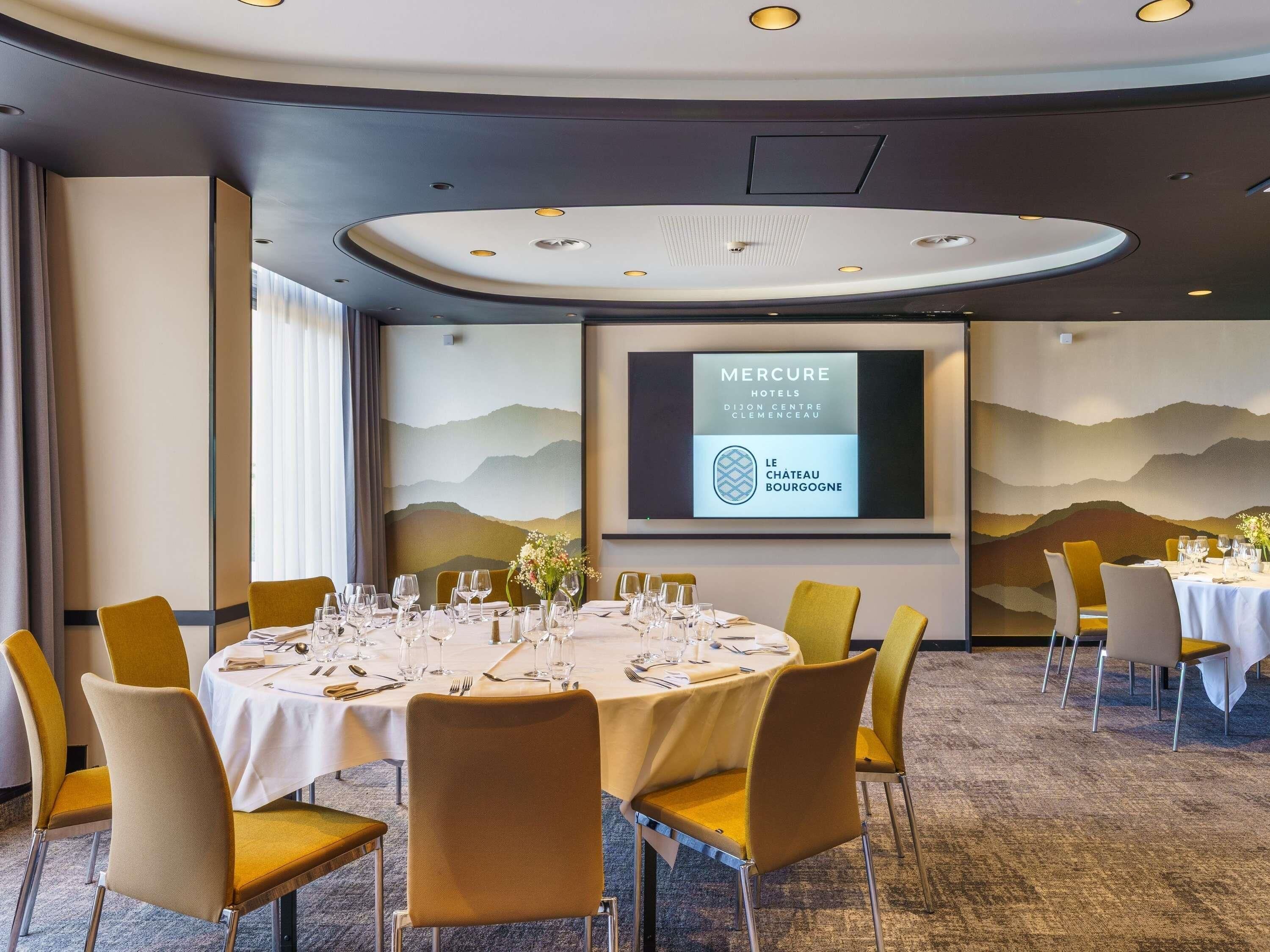 A conference room with a large screen displaying hotel information, round tables with chairs, and a carpeted floor.
