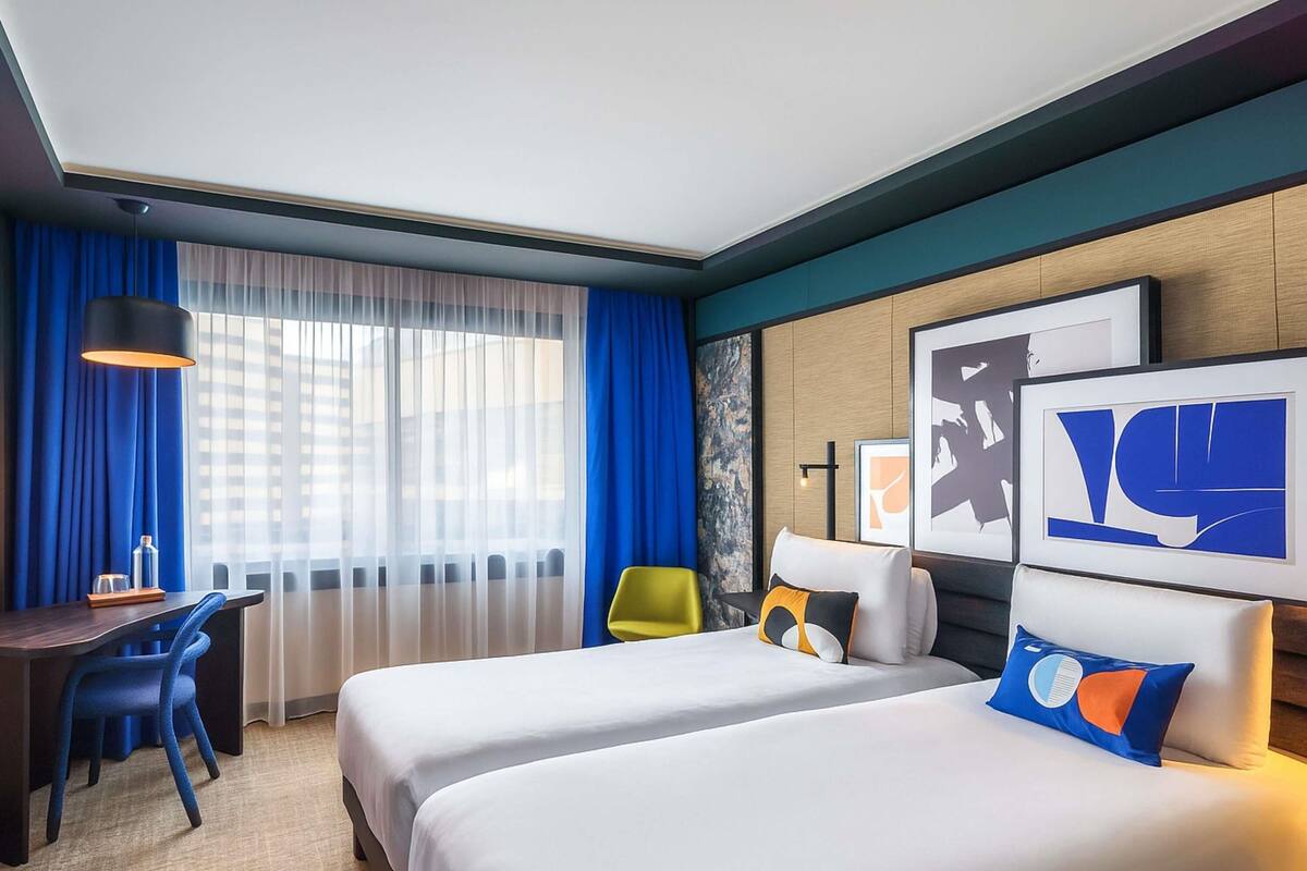 Habitación de hotel con dos camas, un escritorio, una silla y un amplio ventanal con cortinas.