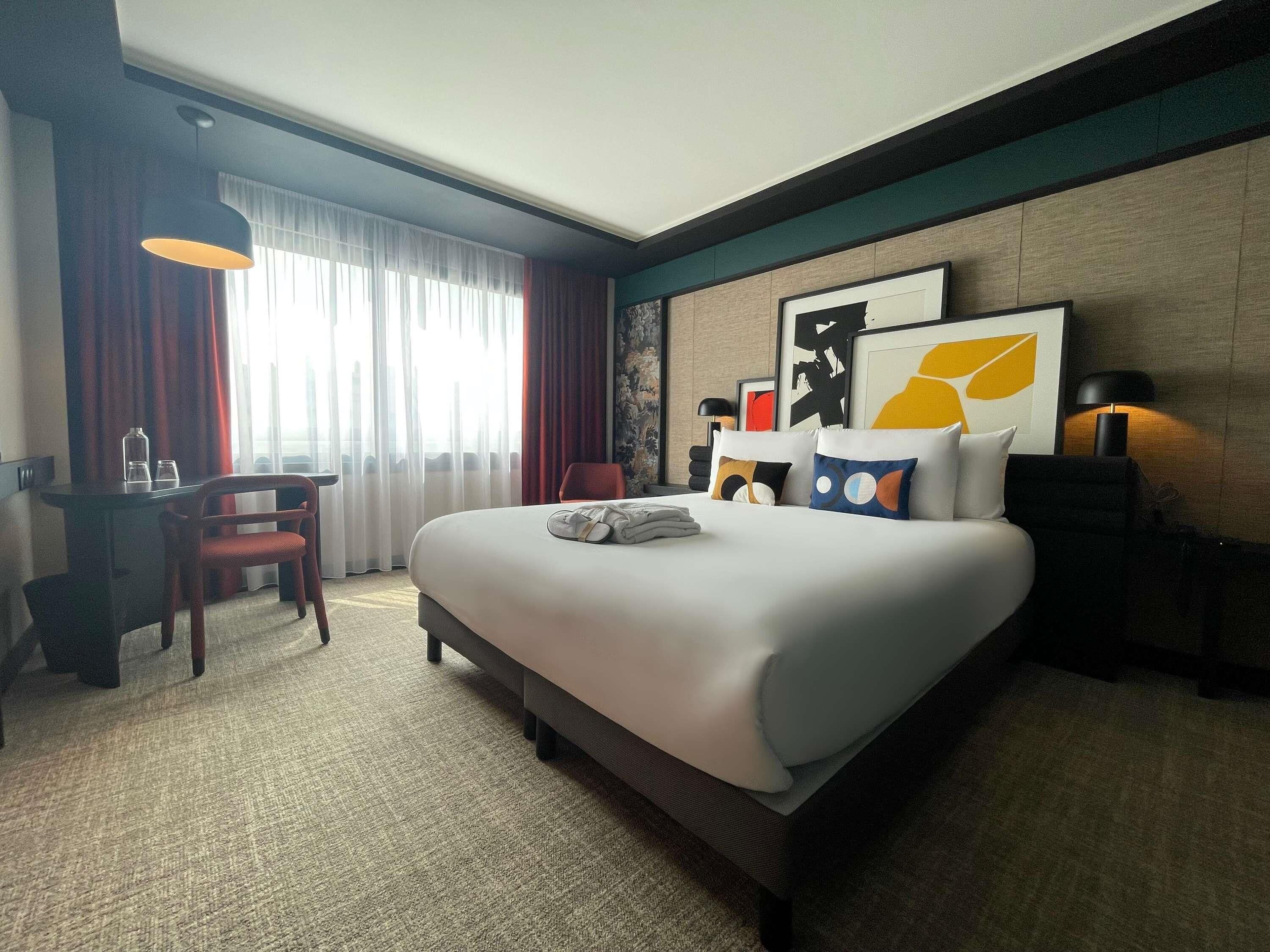 Une chambre d’hôtel avec un grand lit, un bureau avec une chaise, une lampe et des œuvres d’art accrochées au mur.