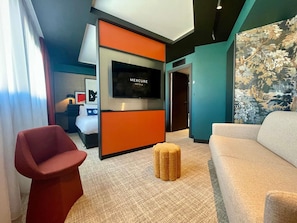 A modern hotel room with a large flat-screen TV, a red chair, a beige sofa, and a patterned wall. - Mercure Dijon Centre Clemenceau (Dijon)
