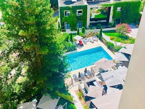 A rooftop pool area with lounge chairs and umbrellas, surrounded by lush greenery and a modern building with a glass facade.