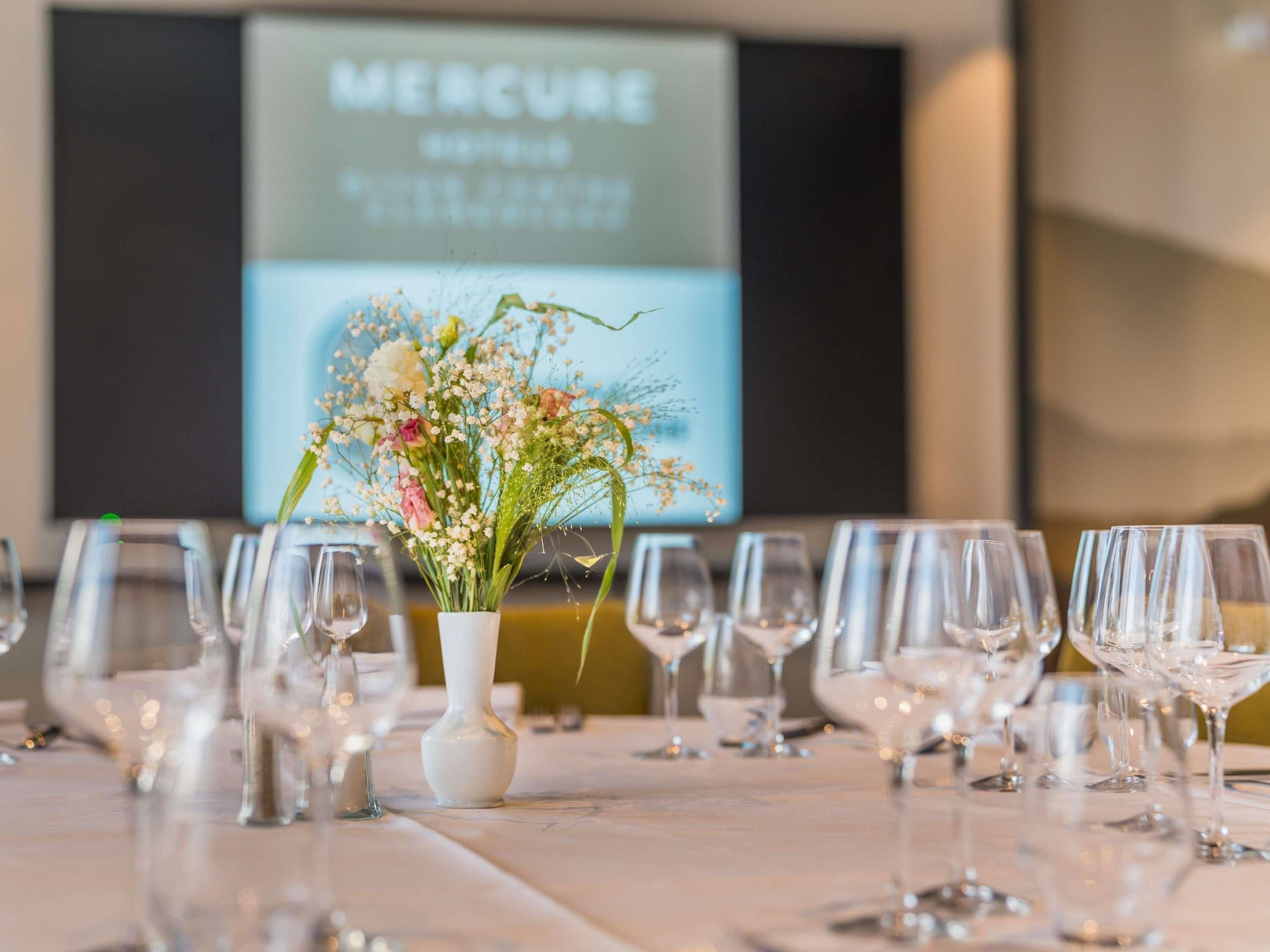 A table set with wine glasses, a white vase with flowers, and a projection screen displaying 'MERCURE' in the background.