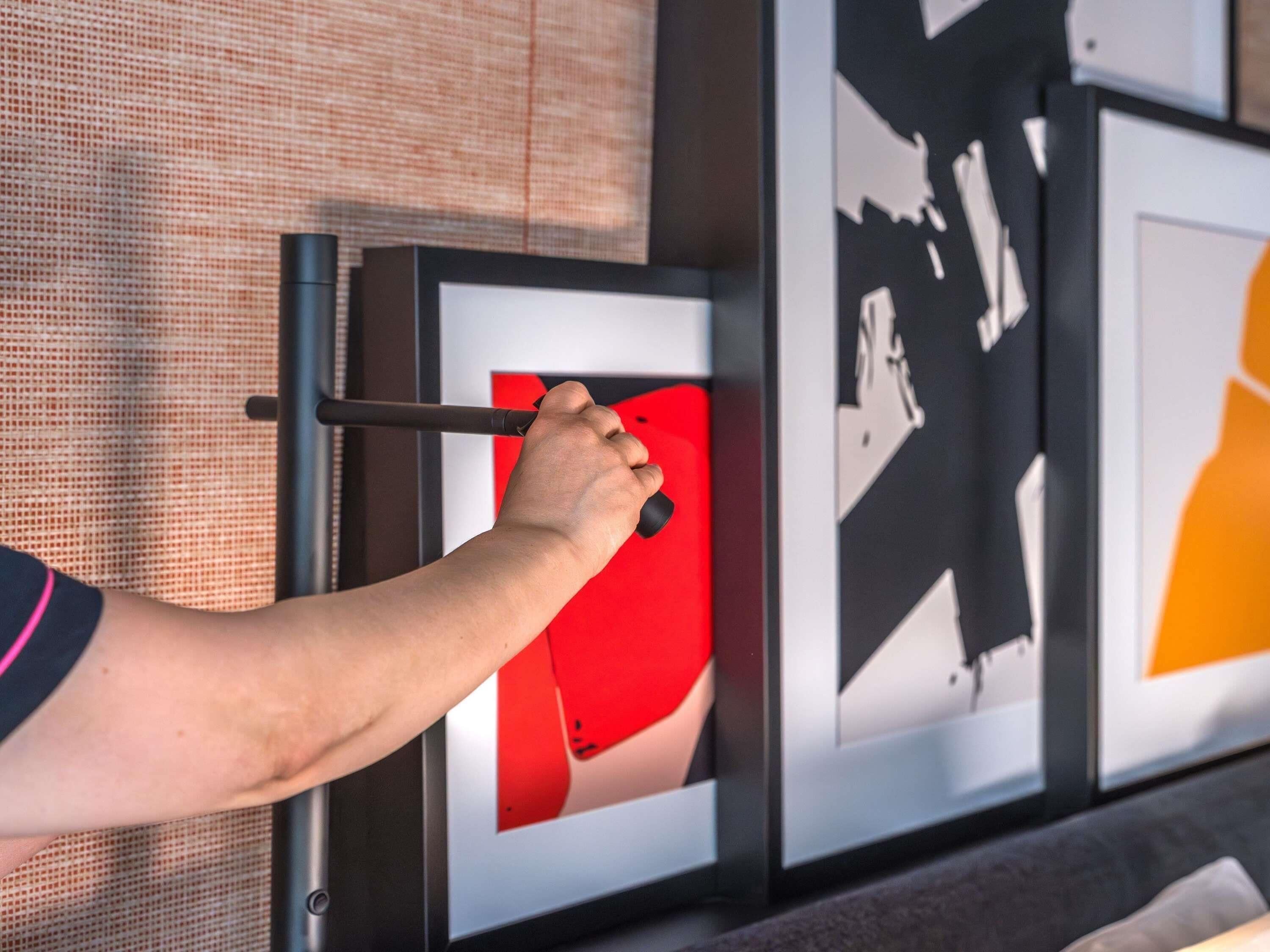 Une main tenant un objet noir, qui pourrait être un outil, près d’une œuvre d’art encadrée dont une partie est rouge.