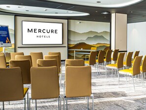 Une salle de conférence avec des rangées de chaises, un grand écran affichant le logo « Mercure Hotels », et une fresque représentant des montagnes.