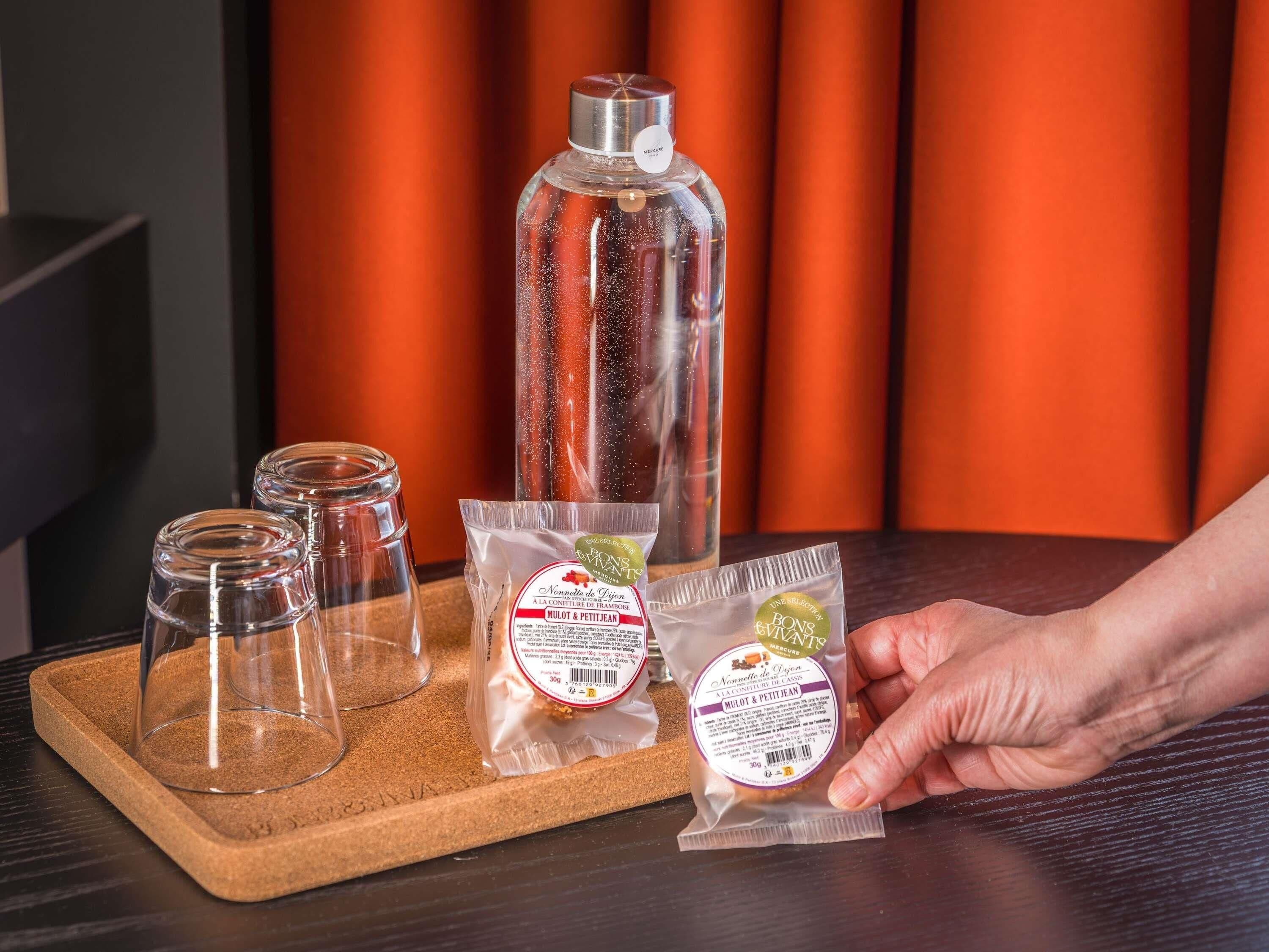 Une main tenant un paquet de biscuits Mullet & Petit Jean, une bouteille en verre et deux verres vides sur un plateau.