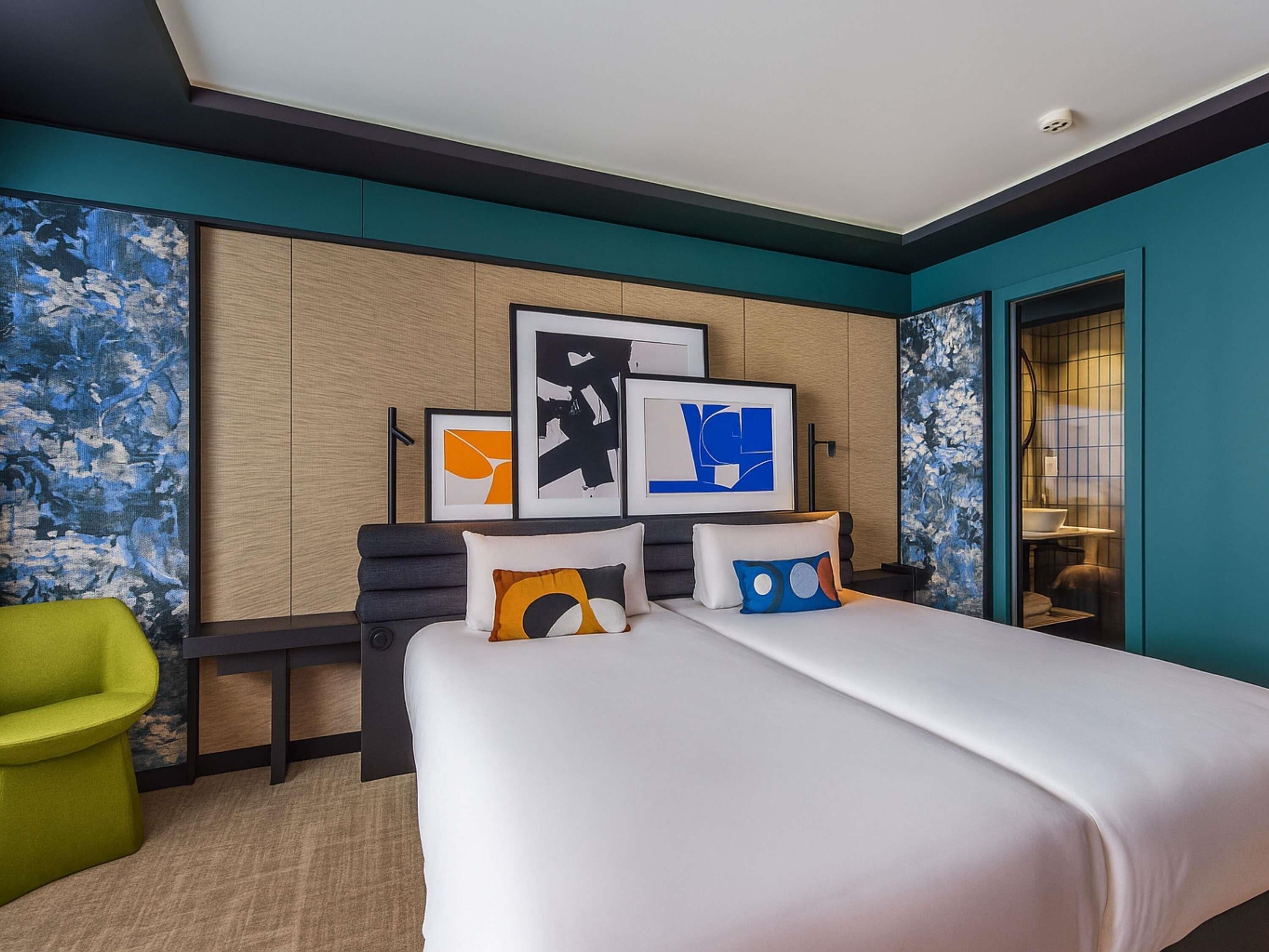 Une chambre d’hôtel avec un lit, un bureau et une chaise. Des œuvres d’art encadrées ornent le mur, et on peut apercevoir la salle de bain à travers une porte ouverte.