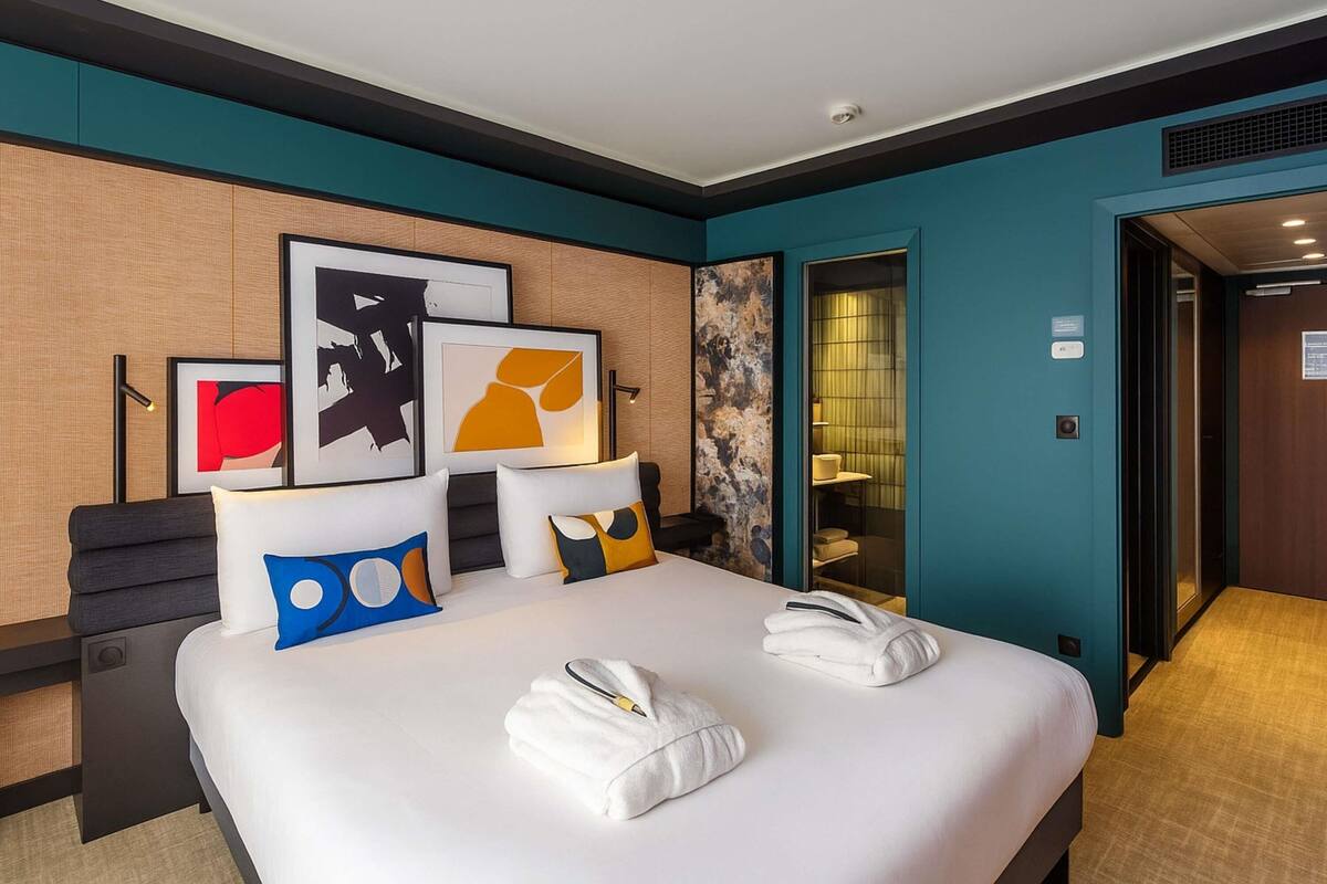 Habitación de hotel con cama, obras de arte, puerta de baño y panel de control.