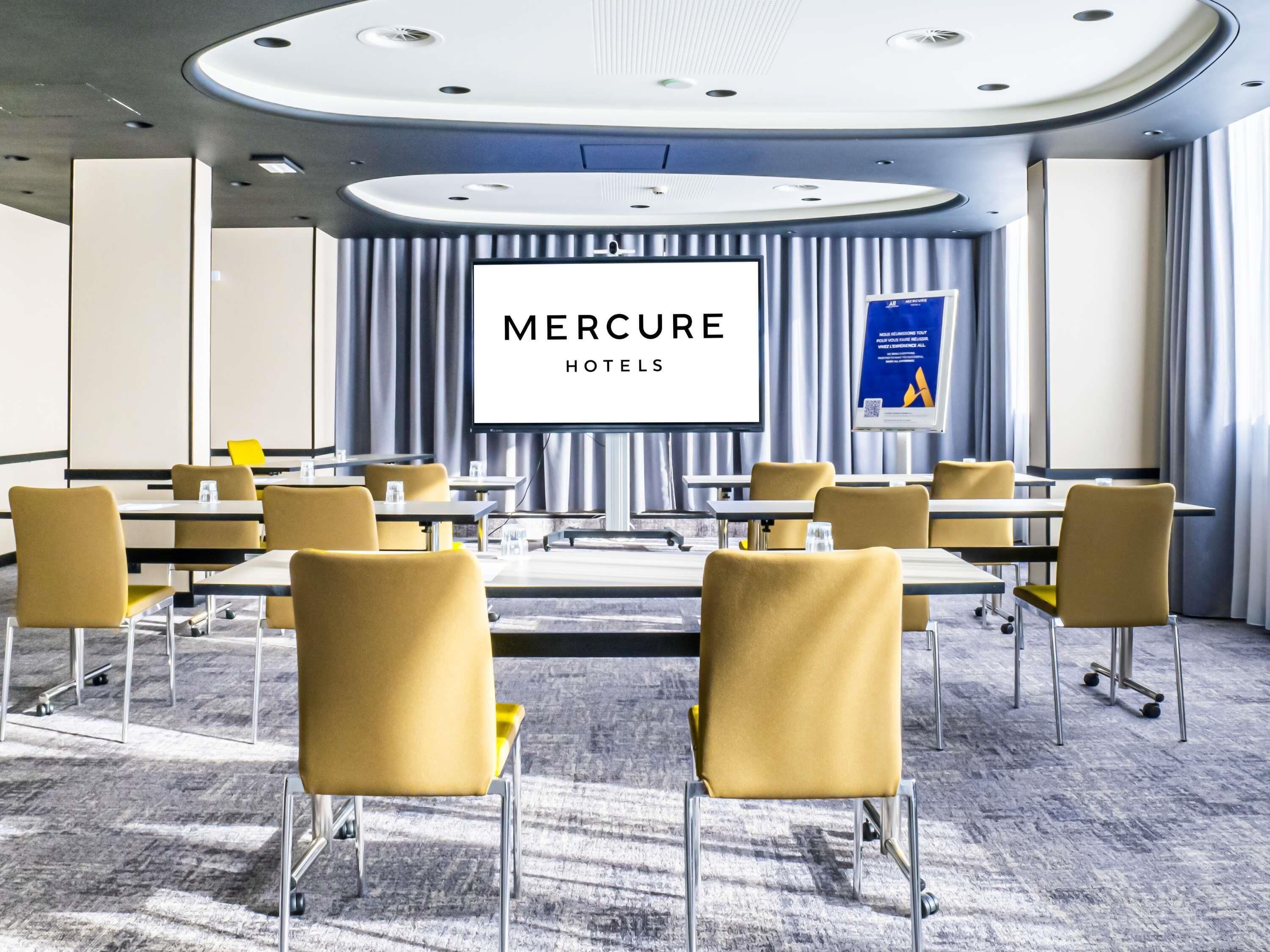 A conference room with a large screen displaying 'Mercure Hotels' branding, multiple tables with chairs, and a carpeted floor.
