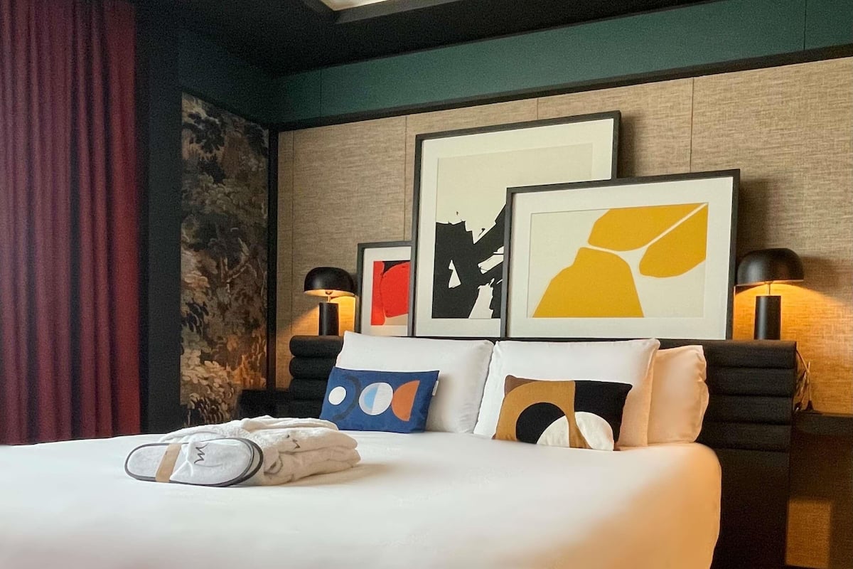 Una habitación de hotel con cama, almohadas, mesita de noche y dos lámparas fijadas a la pared.