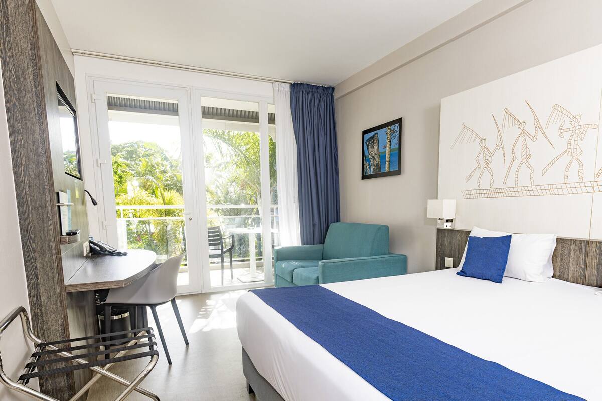 A hotel room with a bed, a desk, a chair, and a balcony with a view of greenery.