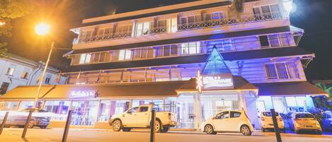 A multi-story hotel with balconies, a well-lit entrance, and parked cars in front.