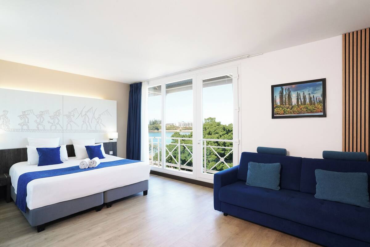 A modern hotel room with a bed, a blue sofa, a balcony with a view, and a painting on the wall.