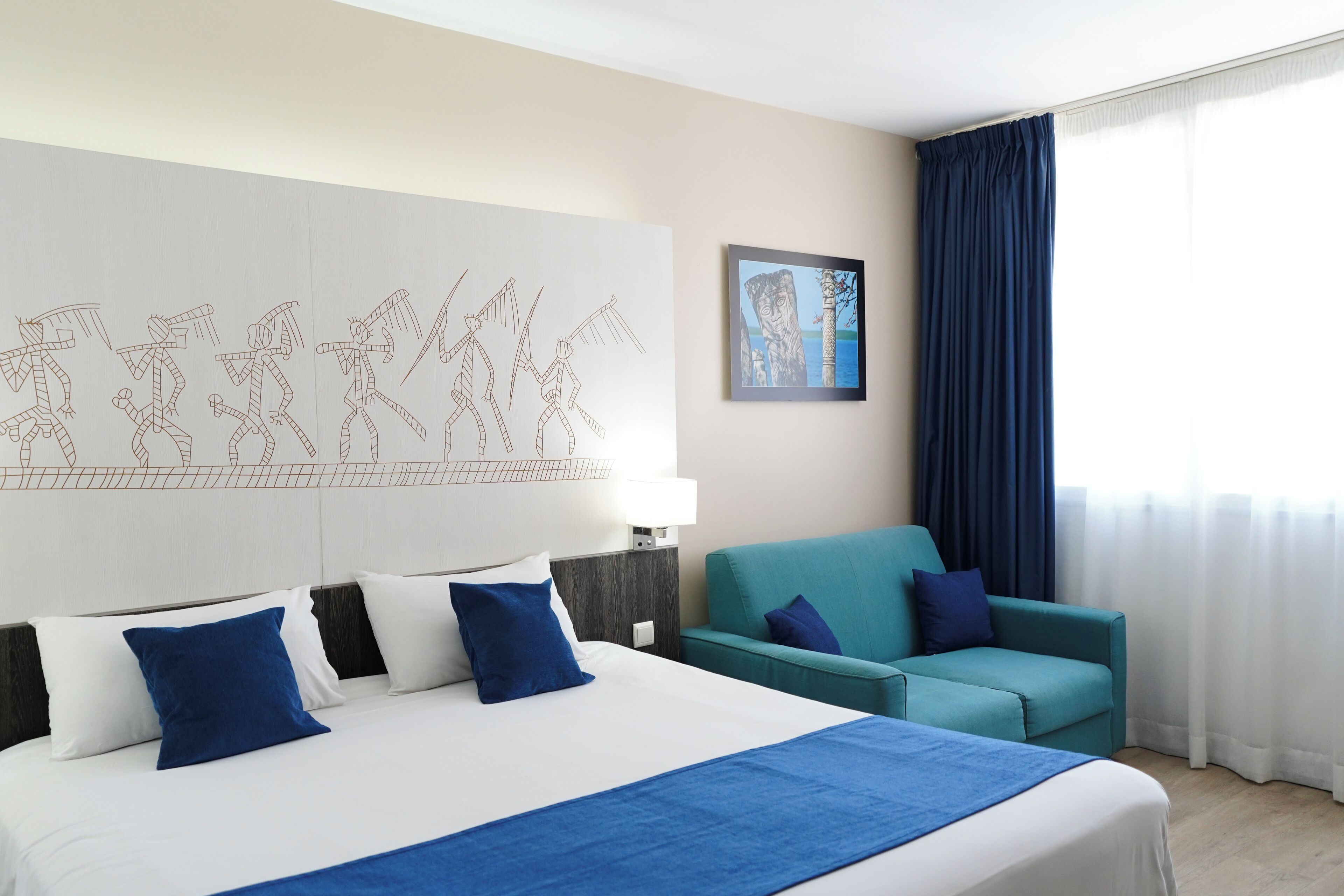 A hotel room with a large bed, a teal sofa, a wall art piece, and a window with curtains.