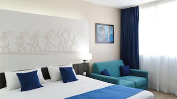 A hotel room with a large bed, a teal sofa, a wall art piece, and a window with curtains.