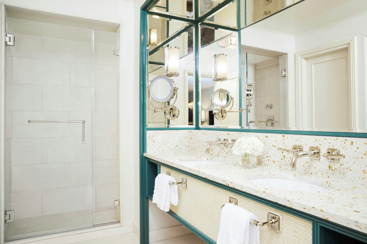 A bathroom with a glass shower enclosure, a large mirror with multiple lights, and a white sink with a towel rack.