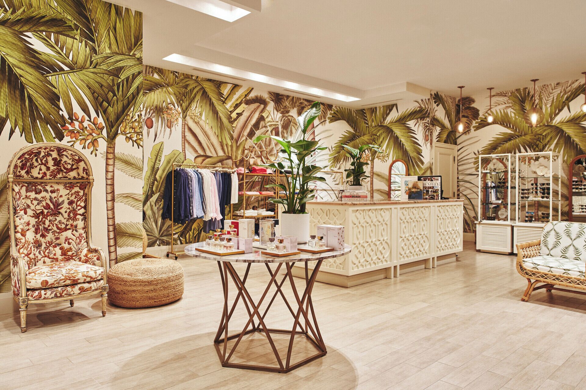 A boutique hotel lobby with a tropical theme, featuring a central round table, a seating area with a patterned sofa, and a display of clothing and accessories.