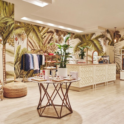 A boutique hotel lobby with a tropical theme, featuring a central round table, a seating area with a patterned sofa, and a display of clothing and accessories.