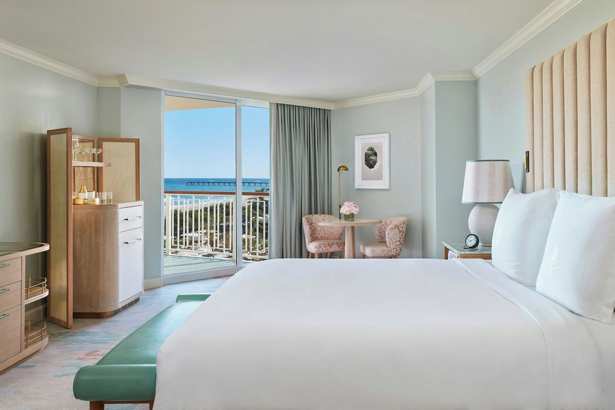 A hotel room with a large bed, a desk with a lamp, a chair, a small table with flowers, and a view of the ocean.